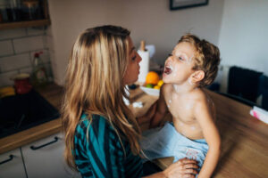 A little boy with measles sits on the kitchen counter. The mother checks if he has a sore throat