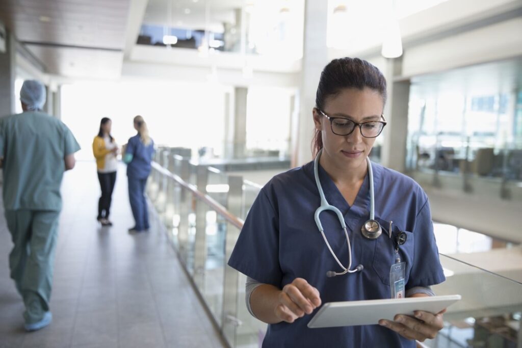 Medical person in scrubs with a stethoscope around their neck is using a tablet device in a walkway with people in the background.